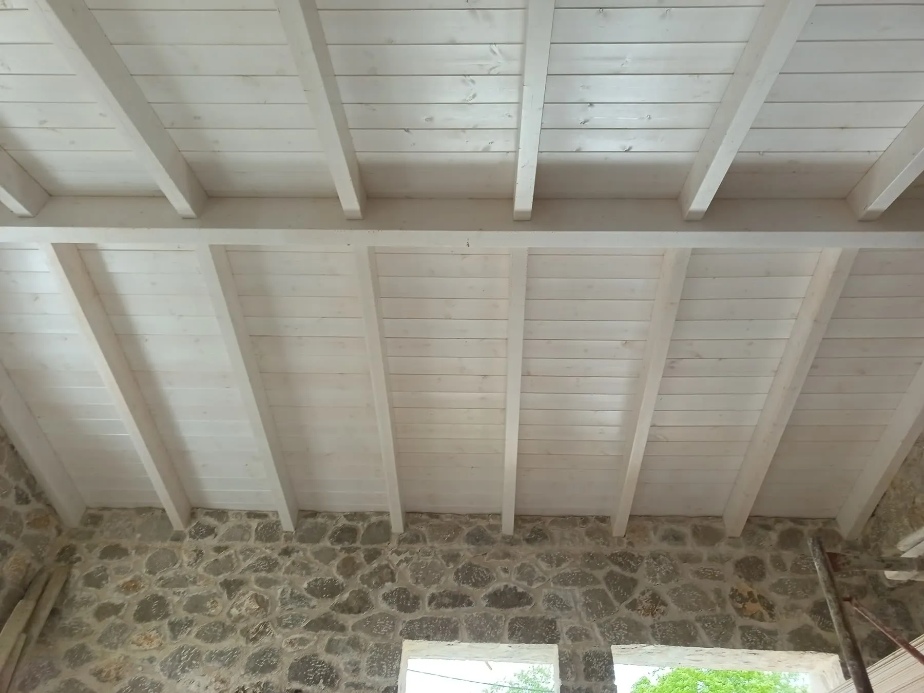A finished timber ceiling seen from below - whitewashed beams and boarding meeting a rough stone wall
