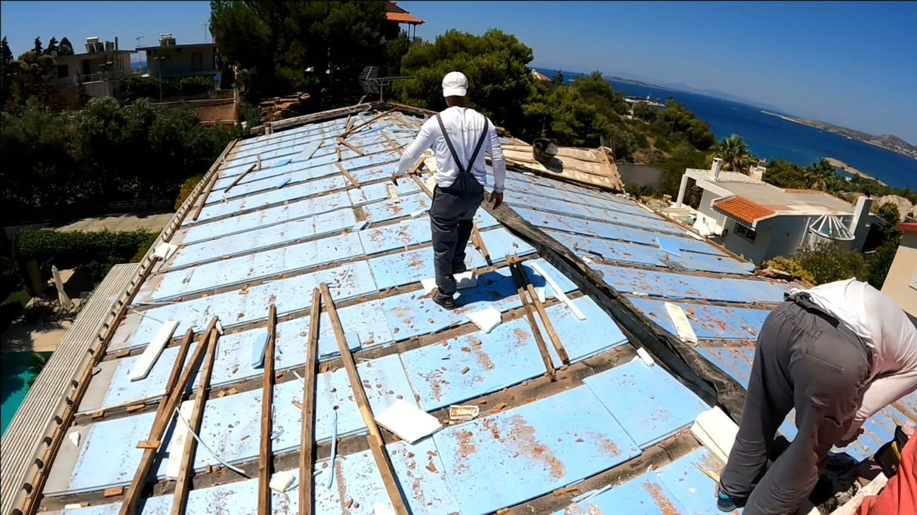 Dismantling phase. The house fully exposed during demolition, Saronic Gulf in the background.