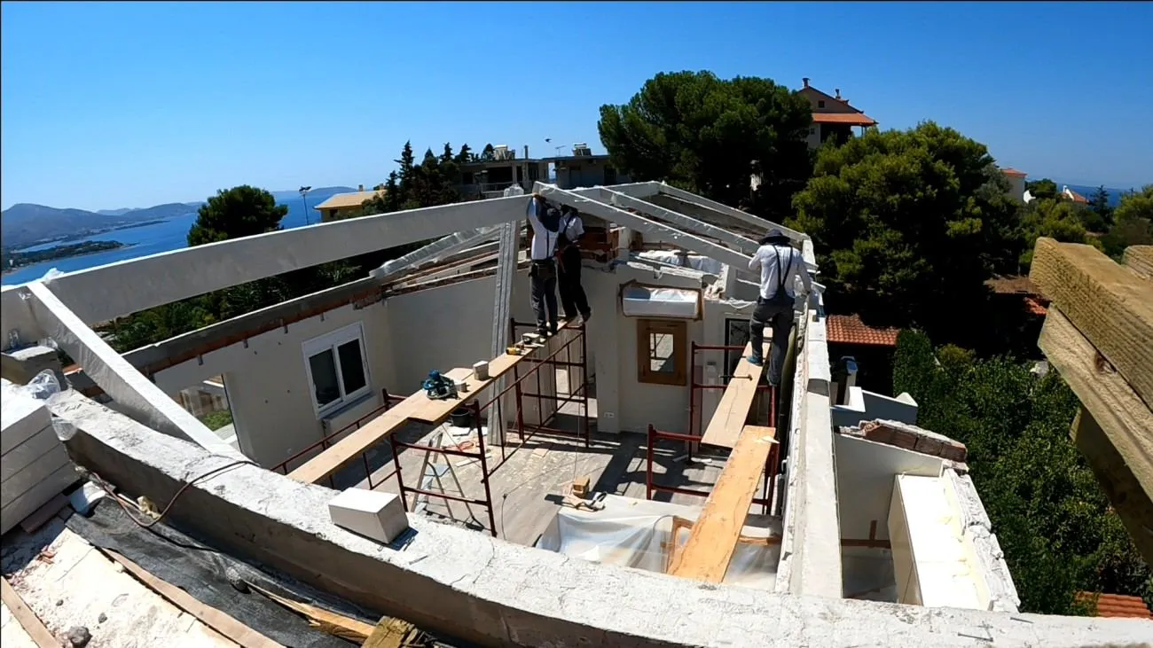 The new skeleton rising. Two workers, the GL24 glulam frame, the sea beyond.